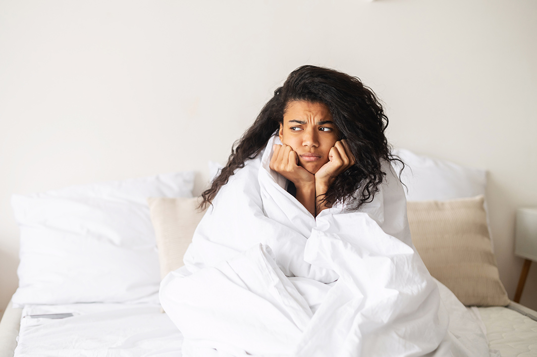 Young African American teenage girl with curly hair placed hands on a chin, covered in a cozy white soft blanket wants to stay at home in the warm bed, feeling suspicious about weather, looking away