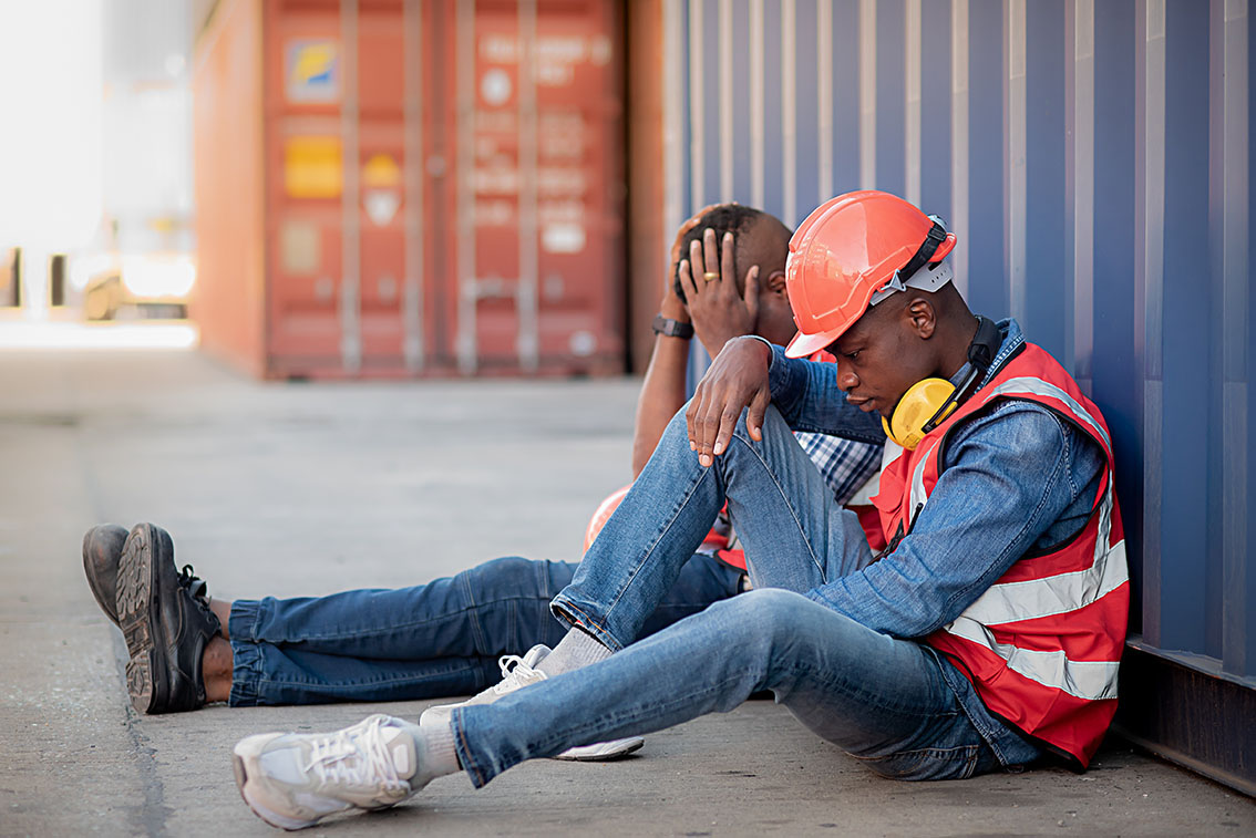 Close-up 2 African American black man worker wearing safety uniform workwear and sitting on the floor leaning against the container because there is no work to do, economic recession due to inflation.