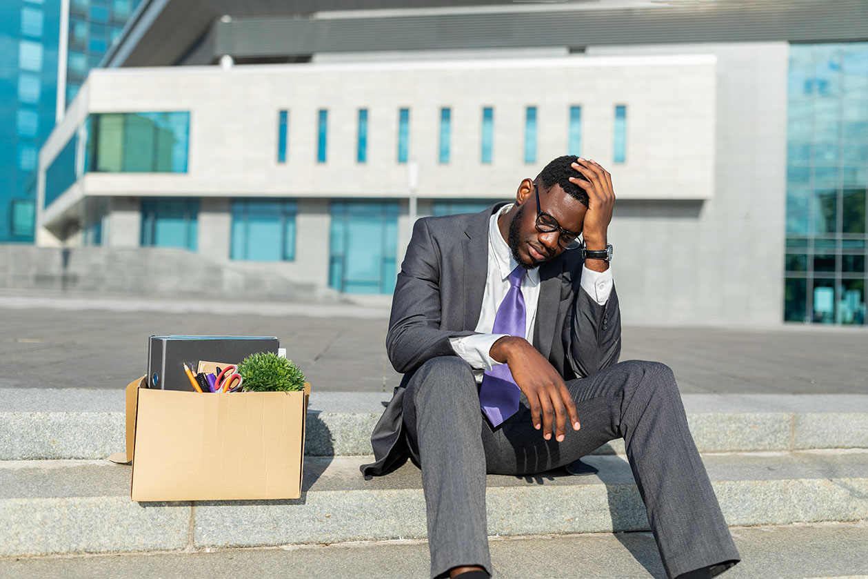 Unemployment concept. Disappointed black businessman sitting with box of personal stuff and empty poster cardboard sign on stairs against office center, got fired and touching head